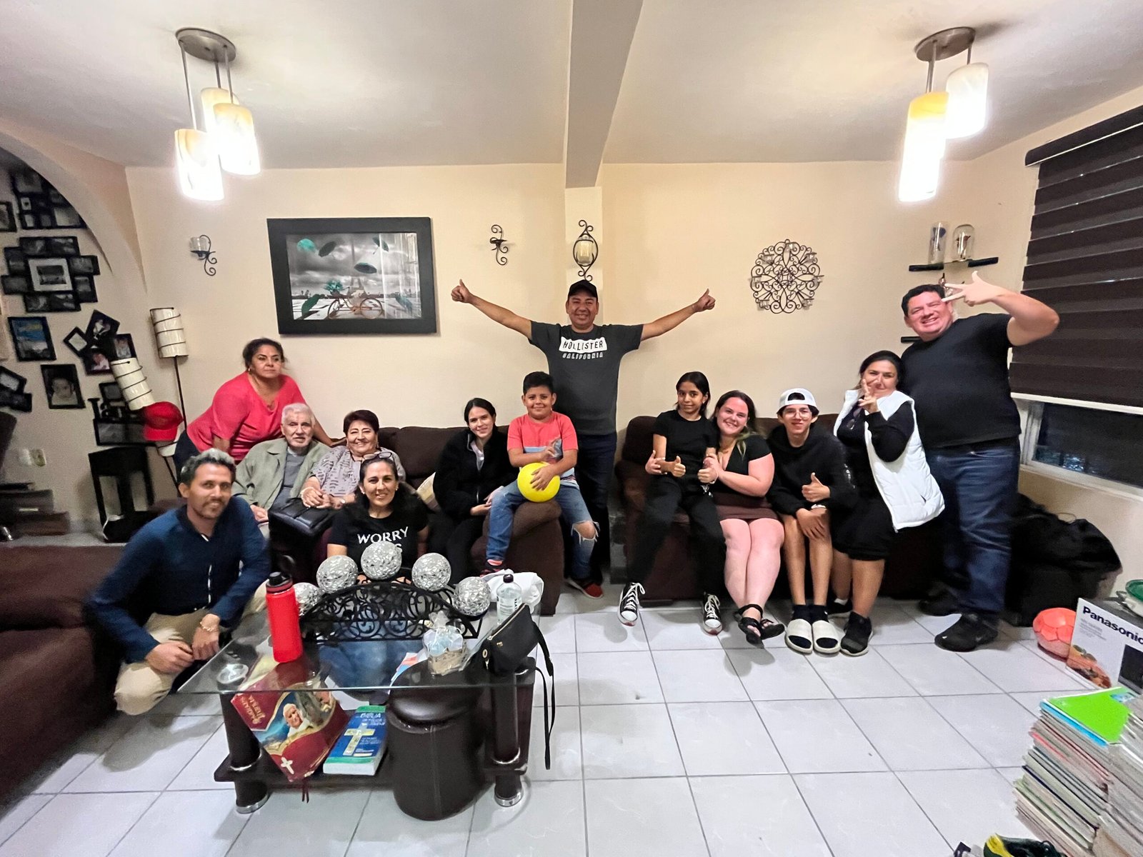 A group of happy adults and kids sit together in a living room with Maestre Paige. They're giving thumbs up and smiling.