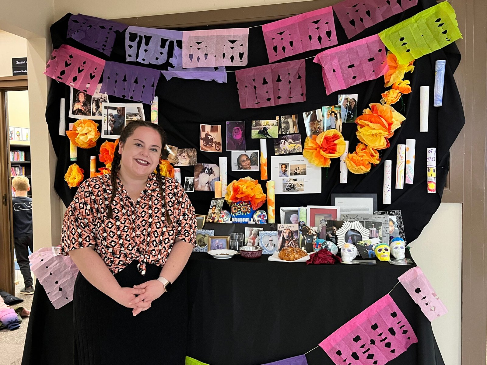 Maestre Paige stands in front of the all-school Day Of The Dead Display which has pictures of lost relatives, friends, and pets amongst offerings of flowers, food, and sugar skulls.