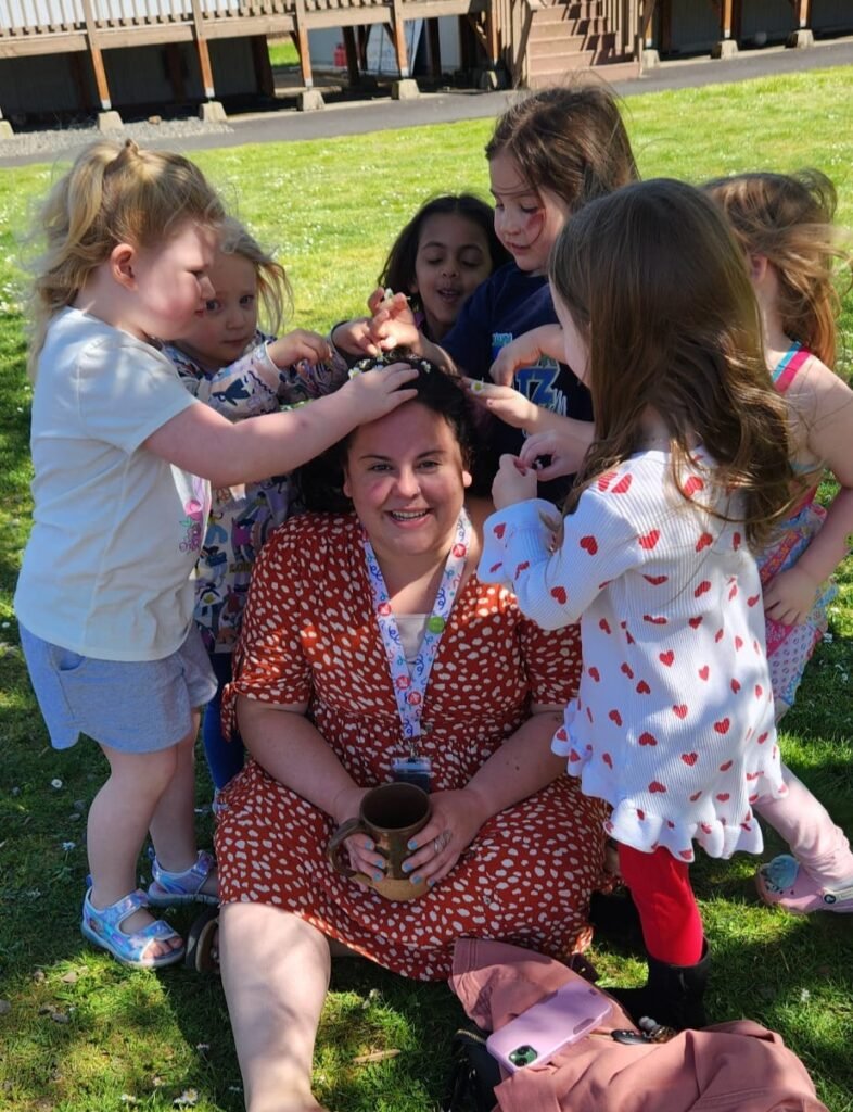 Maestre Paige smiles up at the camera as students surround her, putting daisies in her hair.,