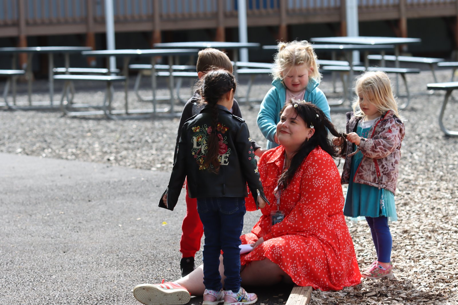 Maestre Paige sits outside with young preschool students. She's smiling up at them as they play with and put flowers in her hair.